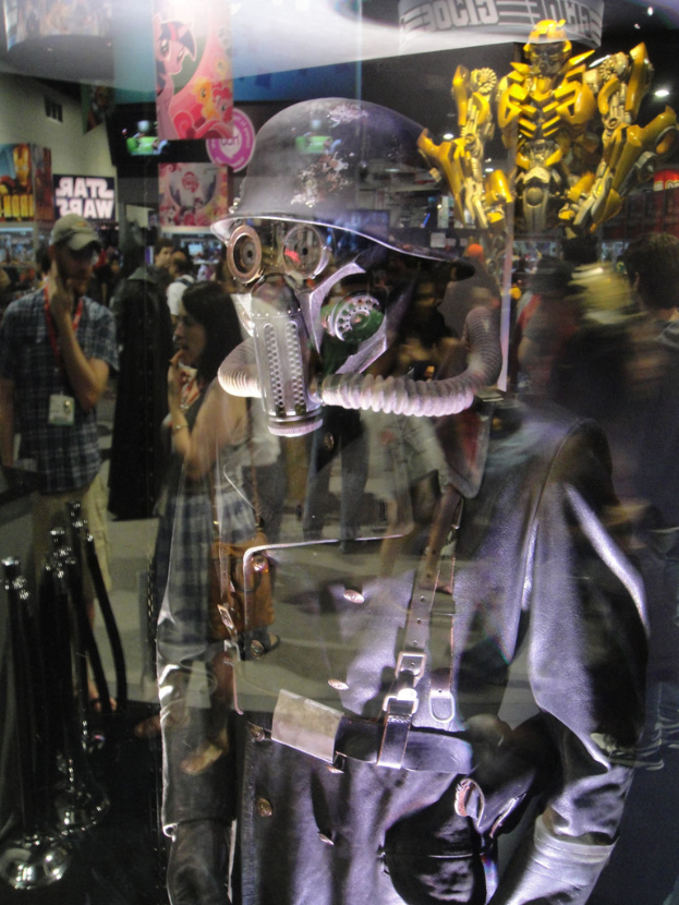 Ein Spiegelbild in einer Halle, das eine Person in Anzug und Helm in der Vordergrund, eine Gruppe von Menschen, Stangen, Poster mit Text, Bilder, eine gelbe Spielzeugfigur und verschiedene andere Gegenstände und Lichter zeigt.