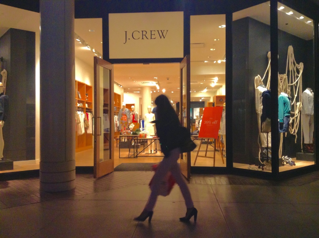 Ein Einkaufszentrum mit Glaswänden und -türen, das Schaufensterpuppen, Tische, hängende Kleider und Schränke sowie eine Frau daneben zeigt.