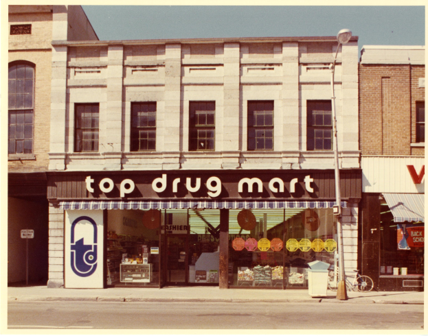 Top-Drogenladen mit einer Straßenecke, einem Gebäude, einem Straßenschild, einem geparkten Fahrrad, einem Müllcontainer und einem bewölkten Himmel.