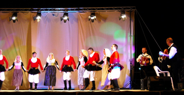 Ausführende auf der Bühne mit einem Vorhanghintergrund, Scheinwerfer oben, Musiker mit Instrumenten rechts und einem weißen Stuhl in der rechten unteren Ecke.