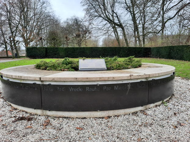Ein Denkmal mit einer Tafel in einem Park, umgeben von Pflanzen, Gras, Bäumen, Häusern, unter einem bewölkten Himmel.