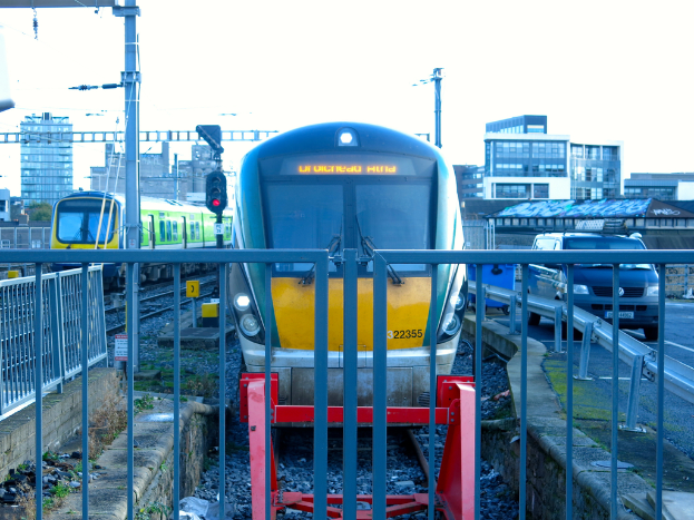 Zwei Züge auf den Schienen mit einem Zaun im Vordergrund, Pfosten auf dem Land, einer mit einer Signalleuchte, ein Auto auf der Straße rechts, Gebäude im Hintergrund und der Himmel oben.