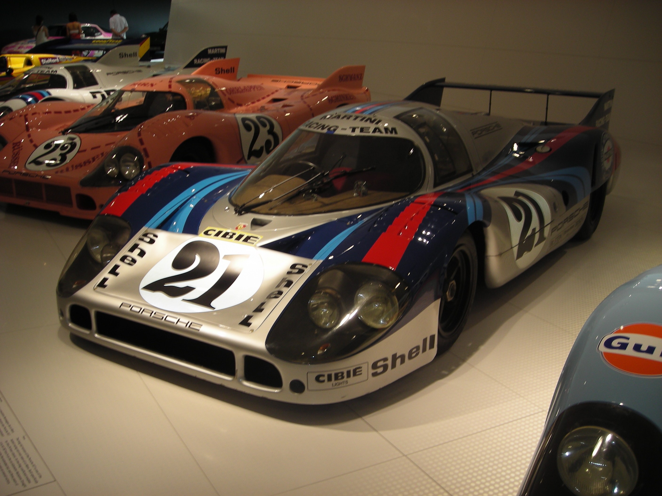 Ein Porsche 917K Rennsportwagen im Museum ausgestellt, umgeben von anderen Sportwagen, mit Menschen in der Nähe und einer Wand im Hintergrund.