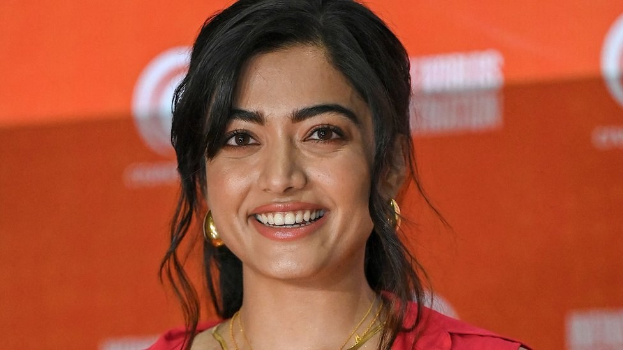 Ein Bild einer Frau in einem roten Kleid mit einer eleganten Hochsteckfrisur und dezentem Make-up, die strahlend lächelt, trägt schwarze High Heels und hat ein hellgelbes Banner mit der Aufschrift 'Die besten Schönheits Looks von Sonam Kapoor beim Cannes Film Festival' im Hintergrund.