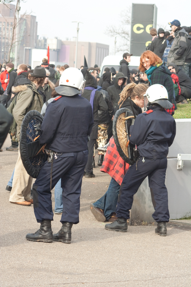 Eine Gruppe von Menschen auf einer Straße mit zwei Personen vorne, die wie Polizisten aussehen, Gebäuden im Hintergrund und Boden unten.