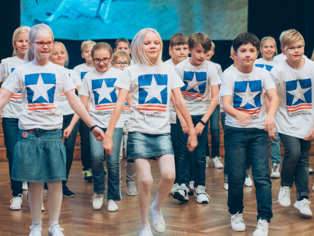 Gruppe von Kindern in weißen Hemden und blauen Shorts, die auf einem Holzfußboden vor einer Wand mit einem Bildschirm tanzen, an einem kunstnädler Tanzwettbewerb teilnehmen.