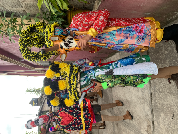 Eine Gruppe von Menschen in bunten Kostümen steht zusammen vor einem Gebäude, mit verschiedenen Gegenständen in der Hand, mit Bäumen, Gebäuden und einem klaren blauen Himmel im Hintergrund während eines Faschingsumzugs.