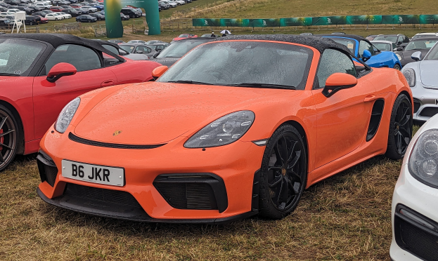 Roter Porsche 718 Boxster GTS auf dem Goodwood Festival of Speed, zusammen mit anderen Autos, einem Banner, Bäumen und einem klaren blauen Himmel im Hintergrund.