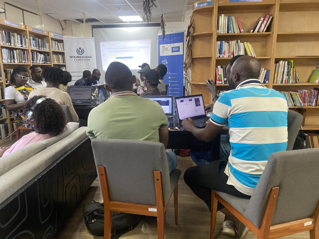 Gruppe von Menschen an einem Tisch mit Laptops, umgeben von Bücherregalen, mit einem Banner, Bildschirm und Deckenbeleuchtung im Hintergrund und Taschen auf dem Boden.