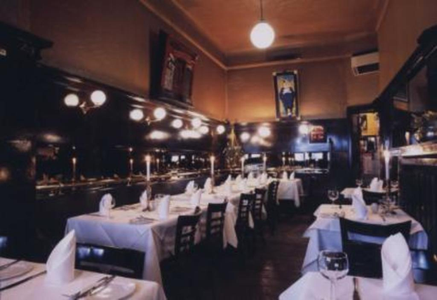 Ein Tisch mit Gläsern, Kerzen in Ständern, Servietten und Tellern, umgeben von Stühlen, mit Lampen und Wandbildern im Hintergrund.