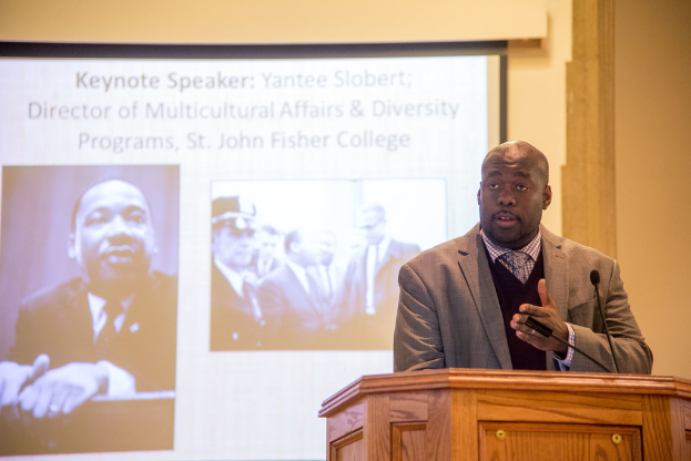 Ein Mann in Anzug, Krawatte und Hemd spricht neben einem Rednerpult auf der rechten Seite des Bildes, mit einem Projektionsschirm in der Mitte und einem Mann in Schwarz-Weiß auf der linken Seite.