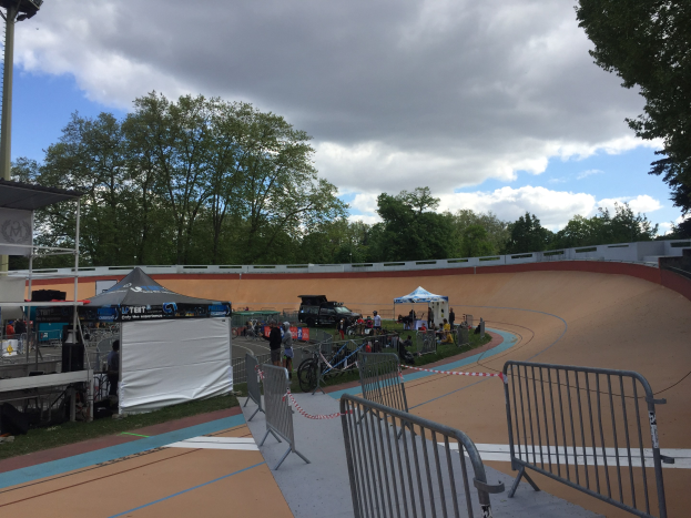 Eine Gruppe von Radfahrern auf einer Schotterstrecke bei den UCI BMX-Weltmeisterschaften 2018, mit Zelten, Fahrzeugen, Gittern, Bäumen und einem bewölkten Himmel im Hintergrund.