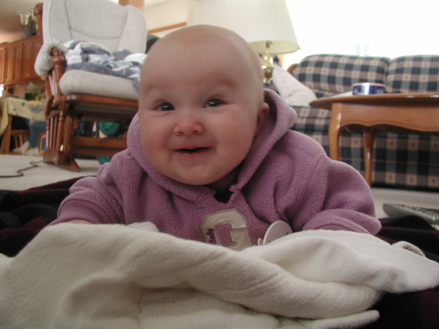 Ein Junge lächelt in einem Haus mit einem Stuhl, einer Couch, einem Zentertisch, einer Lampe, einem Kleiderschrank und einem Tisch im Hintergrund, mit einem Kabel und Decken auf dem Boden.
