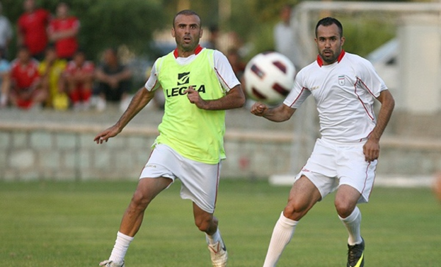 Zwei Spieler rennen auf einem Rasenfeld mit einem Ball in der Luft, während Zuschauer und Bäume im Hintergrund sichtbar sind.