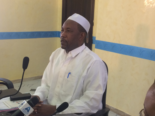 Ein Mann in einem weißen Kleid und einer Mütze sitzt an einem Tisch mit Mikrofonen, spricht, während Papiere und Gegenstände auf dem Tisch zu sehen sind, mit einer anderen Person mit Brille rechts von ihm, in einem Raum mit einer Wand und einer Tür im Hintergrund.