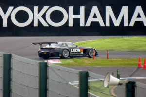 Schwarzer Sportwagen auf einer Rennstrecke mit Verkehrszeichen, Gras und einem Zaun, mit einem "Yokohama"-Schild im Hintergrund und einem Lautsprecher im Vordergrund.