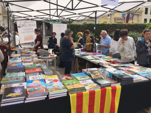 Eine Gruppe von Menschen auf einer Buchmesse in Barcelona, die um einen mit einem Tuch bedeckten und mit Büchern gefüllten Tisch stehen und einige Bücher in der Hand halten, mit einem Banner, Pflanzen und Gebäuden mit Fenstern im Hintergrund unter einem klaren blauen Himmel.