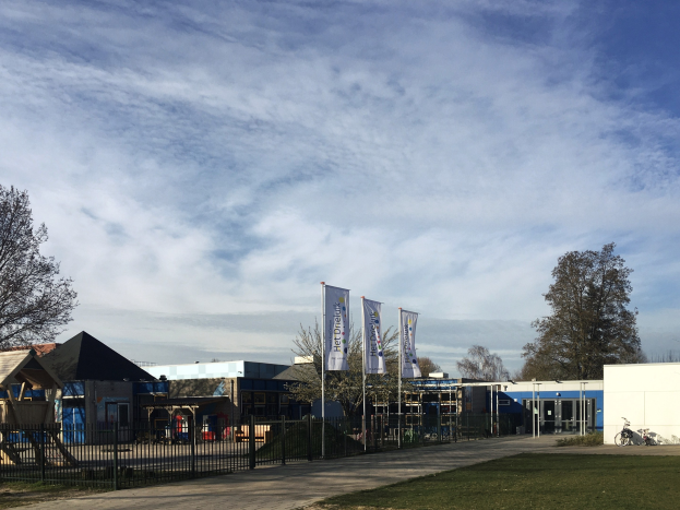 Großes Gebäude mit einem großen Rasen vorne, umgeben von einem Metallzaun, Bäumen, Bannern, einem Fahrrad, nahen Häusern und einem bewölkten Himmel, als neuer Spielplatz für Kinder identifiziert.