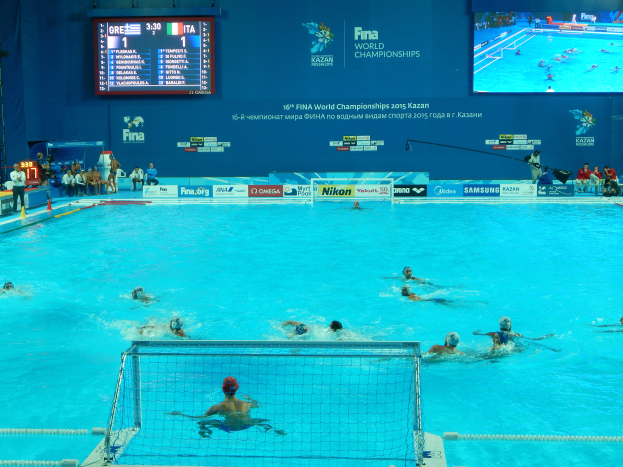 Eine Gruppe von Menschen spielt Wasserball in einem großen Pool mit einem Netz in der Mitte, Zuschauern auf der linken Seite und einem Bildschirm mit Text und Logos im Hintergrund bei den 2016 FINA World Championships in Kazan.