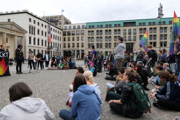 Eine Gruppe von Menschen, die auf dem Boden vor einer Menge sitzt, die Fahnen und Schilder hält, mit einer Person, die in ein Mikrofon spricht, einer Statue und Gebäuden im Hintergrund während einer anti-schwulen Demonstration in Berlin, Deutschland.