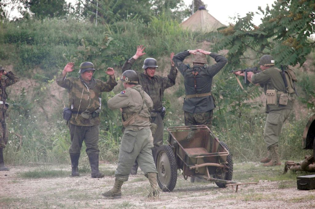Mehrere Personen in militärischer Uniform und Helmen stehen in der Nähe eines Wagens, umgeben von Pflanzen, Bäumen und Gras unter einem klaren Himmel.