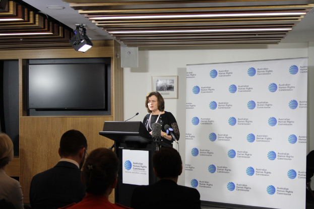 Eine Frau steht auf einer Bühne und spricht in ein Mikrofon, vor ihr sitzt ein Publikum, an der Wand hängt ein Banner, auf dem Holzfußboden steht ein Fernseher und an der Decke leuchten Lampen.