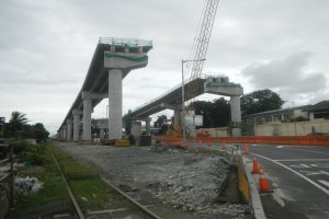 Baustelle mit einer Brücke im Hintergrund, eine Straße mit Absperrkegeln auf der rechten Seite, Steine und Gras am Boden, eine Bahnschiene auf der linken Seite, Bäume und Gebäude auf beiden Seiten der Straße und ein bewölkter Himmel.