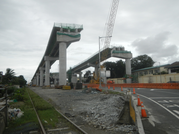 Baustelle mit einer Brücke im Hintergrund, eine Straße mit Absperrkegeln auf der rechten Seite, Steine und Gras am Boden, eine Bahnschiene auf der linken Seite, Bäume und Gebäude auf beiden Seiten der Straße und ein bewölkter Himmel.
