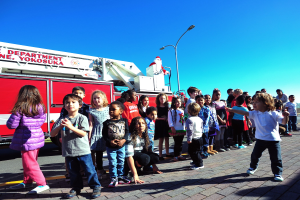 Eine Gruppe von Kindern vor einem Feuerlöschwagen auf einer Straße mit Laternenpfählen, Bäumen und einem klaren blauen Himmel, der Feuerlöschwagen zeigt Texte im Zusammenhang mit der Weihnachtsparade der Yokosuka-Feuerwehr.