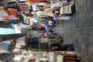 Ein belebter Straßenmarkt in der Altstadt von Jerusalem, mit Menschen, die an Ständen mit Kleidung, Taschen, Lampen und anderen Artikeln stöbern, und einem Gebäude im Hintergrund.