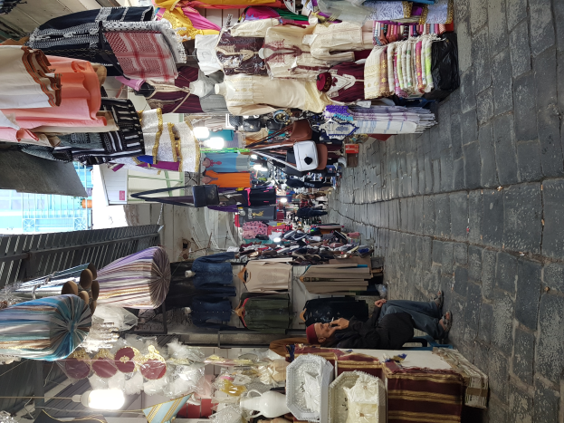 Ein belebter Straßenmarkt in der Altstadt von Jerusalem, mit Menschen, die an Ständen mit Kleidung, Taschen, Lampen und anderen Artikeln stöbern, und einem Gebäude im Hintergrund.