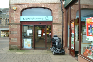 Apothekenladen mit einem Fahrzeug davor und einem Gebäude links daneben.