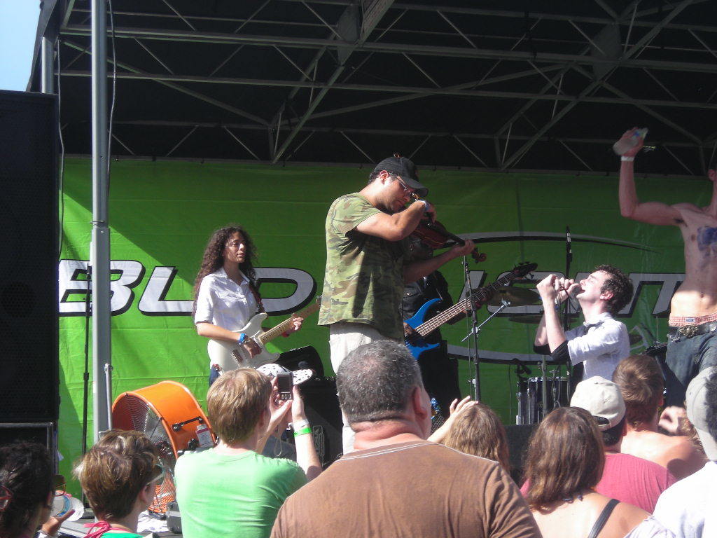 Eine Gruppe von Musikern, die verschiedene Instrumente auf einer Bühne spielen, während das Publikum davor steht und genießt, mit einem grünen Banner im Hintergrund.