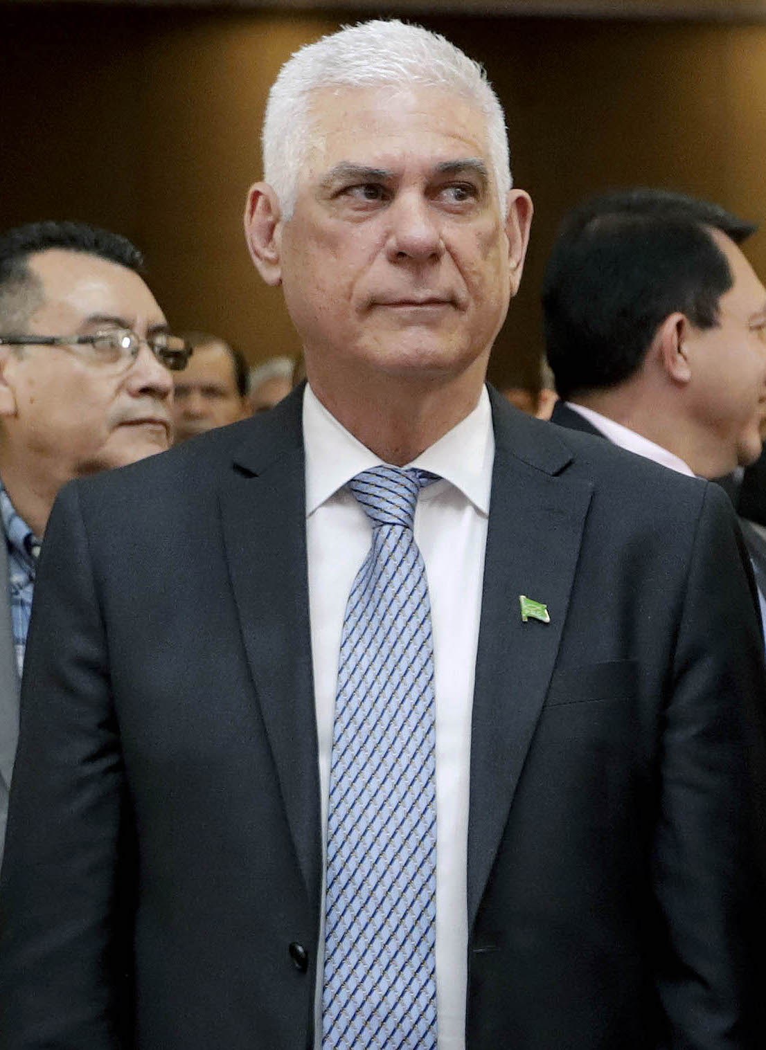 Ein Mann in einem blauen Anzug, weißem Hemd und gemusterter Krawatte steht vor einer Gruppe von Menschen und spricht mit ernster Miene.