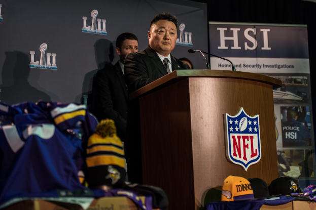 Ein Mann in einem schwarzen Blazer, einem wei├čem Hemd und einer schwarzen Krawatte spricht an einem Pult mit einem Mikrofon und Logo vor einer Menge, mit blauer und gelber Merchandise zu seiner Linken und einem "Super Bowl LII"-Banner im Hintergrund.