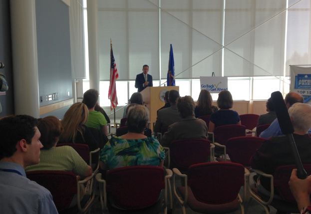 Ein Mann steht an einem Rednerpult und spricht zu einem Publikum, das auf Stühlen sitzt, mit zwei Fahnen neben ihm und einer Wand, Säulen und Fenstern im Hintergrund.