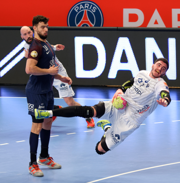 Eine Gruppe von M├Ąnnern, die Handball auf einem Platz spielen, einer mit einem Ball, mit einem Hintergrundplakat der Paris Saint-Germain Futsal-Mannschaft.