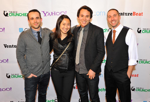 Eine Gruppe lachender Menschen posiert vor der Kamera mit einem Sponsorenbanner im Hintergrund.