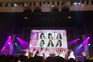 Eine Gruppe von Menschen performs auf einer hell erleuchteten Bühne mit Trägern, während eine Person ein Mikrofon hält und eine andere ein Instrument spielt; eine Menge unten zeichnet das Ereignis auf, und ein großer Bildschirm im Hintergrund zeigt Bilder der K-Pop-Gruppe BK48.