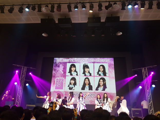 Eine Gruppe von Menschen performs auf einer hell erleuchteten Bühne mit Trägern, während eine Person ein Mikrofon hält und eine andere ein Instrument spielt; eine Menge unten zeichnet das Ereignis auf, und ein großer Bildschirm im Hintergrund zeigt Bilder der K-Pop-Gruppe BK48.