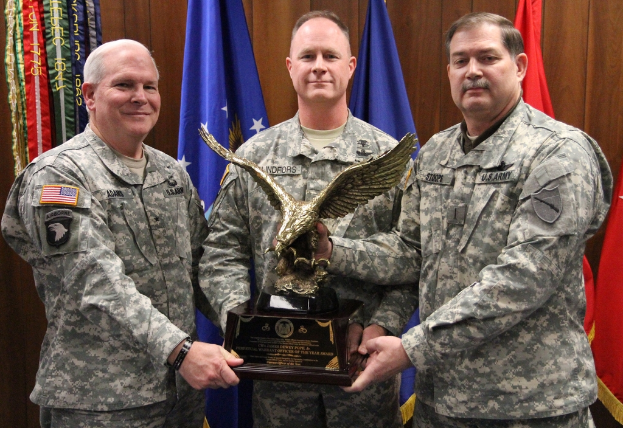 Drei Männer in Uniformen halten Auszeichnungen, mit Flaggen im Hintergrund und buntem Text in der oberen linken Ecke.
