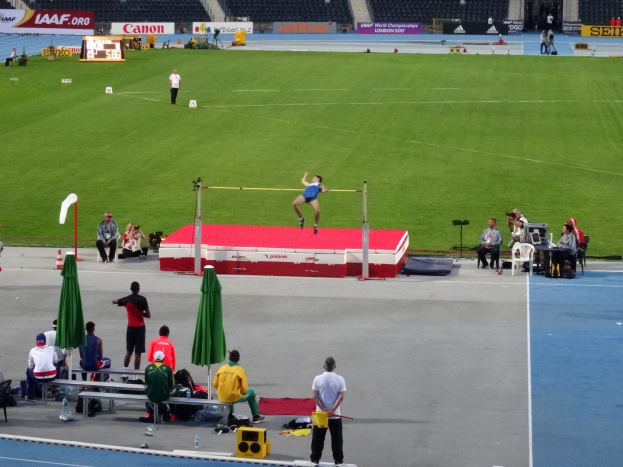 Ein Mann springt über eine Hochsprungstange auf einer Laufbahn, mit Zuschauern im Hintergrund, Schirmchen, Verkehrskegel und Texttafeln.