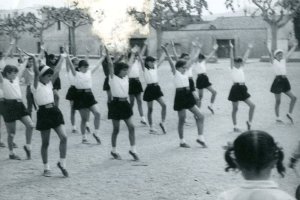 Schwarzes und weißes Foto von jungen Mädchen in Schuluniformen, die auf einer von Gebäuden und Bäumen gesäumten Straße tanzen, mit einer Person in Schuhen unten im Bild.