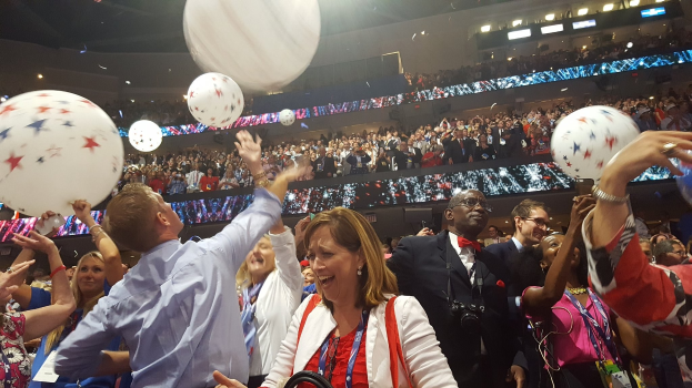 Eine Menschenmenge steht im Freien, einige halten Luftballons, während andere im Hintergrund auf Stühlen sitzen, unter beleuchteten Lichtern, die scheinbar einen Sieg bei der Republican National Convention feiern.