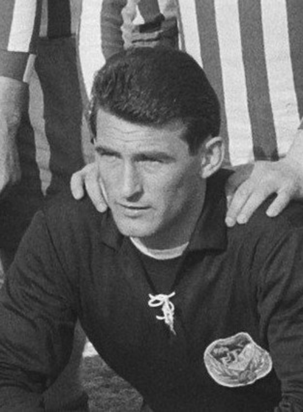 Schwarzes und weißes Foto eines Mannes, der auf einem grünen Fussballfeld sitzend ein T-Shirt und eine Kette trägt, mit zwei Personen dahinter.