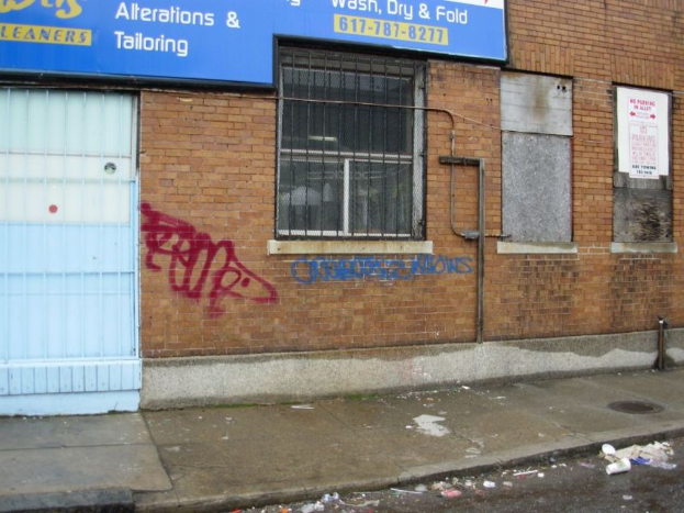 Eine Straßenszene mit einem Fußweg und einer Straße, die mit Müll übersät sind, eine Backsteinmauer, Fenster, Rohre, ein Tor und eine Wand am oberen Rand.