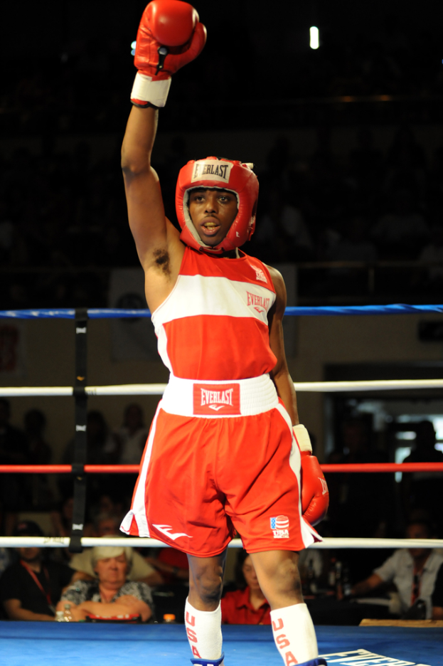 Ein Mann in Boxerausrüstung, einschließlich Helm und Handschuhen, steht in einem Boxring mit Zuschauern im Hintergrund.