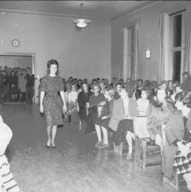 Eine Frau in einem schwarzen und weißen Kleid läuft über einen Laufsteg während einer Modenschau für Frauenrechte, mit einer Menge, die auf Stühlen sitzt und einer Uhr und Lichtern an der umliegenden Wand.