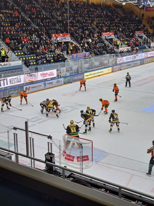Eine Gruppe von Menschen, die Hockey auf einem Eisstadion spielt, mit einem Netz, einer Umrandung, sitzenden Zuschauern und beleuchteten Lichtern.