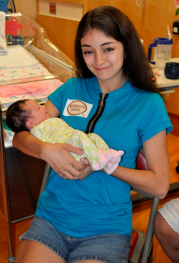 Eine Frau in einem blauen T-Shirt sitzt auf einem Stuhl und lächelt, während sie ein Baby mit beiden Händen hält, mit einem Bett, einem Krug und einem Gegenstand im Hintergrund auf einem Tisch.